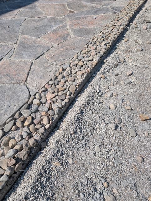 Hand-fit granite flagstone with tight swept joints, edged by a band of washed river stone against the gravel drive. — Collingwood, Ontario