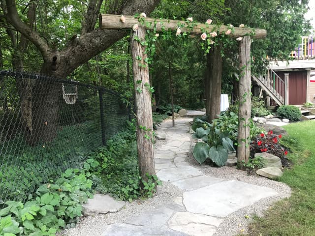 Peeled log post-and-beam arbour set over an irregular flagstone walkway, with climbing vine trained up the posts and the path edged in stone against pea gravel. — Barrie, Ontario