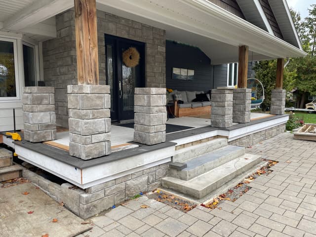 Front porch with stone-clad columns and dark stone caps, poured concrete steps set into the paver patio.